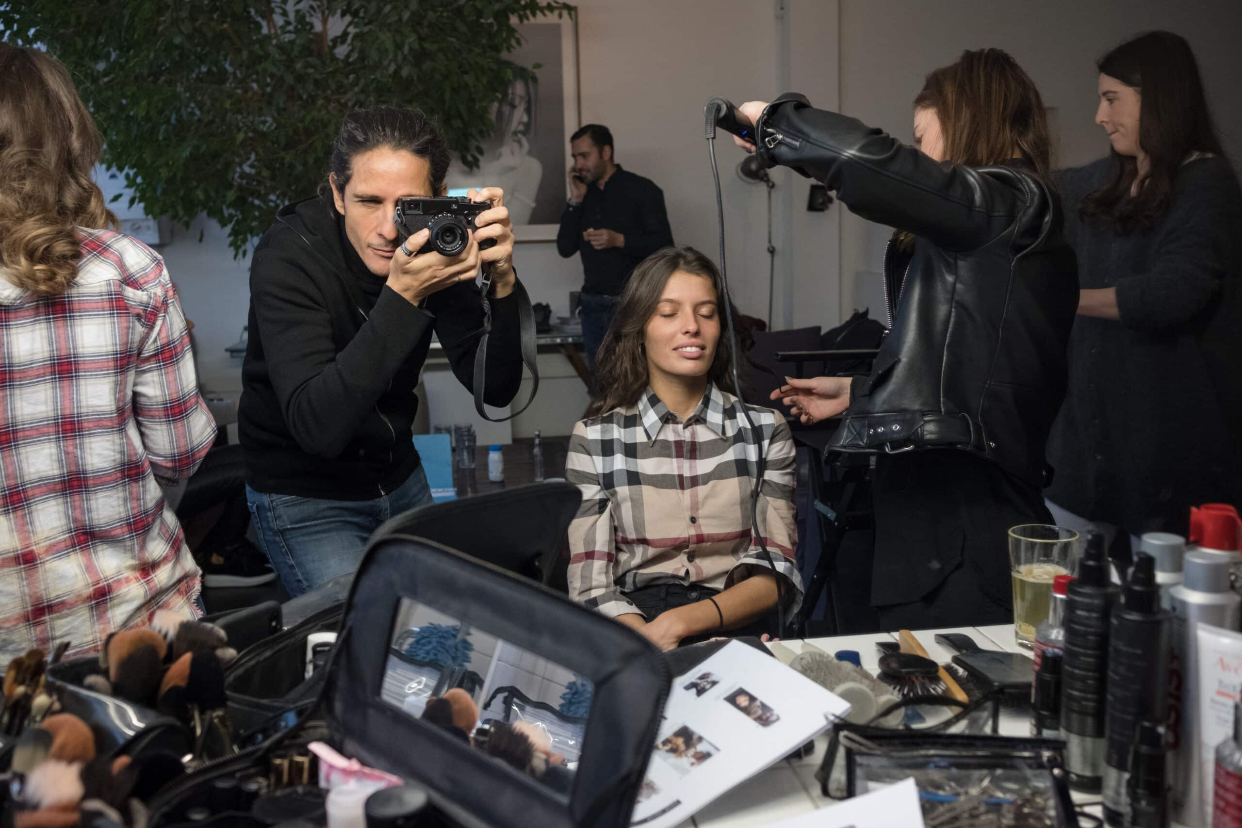 Vue des coulisses d'un shooting de mode montrant un photographe prenant une photo d'un mannequin assis, pendant qu'une coiffeuse s'occupe de ses cheveux. Au premier plan, une table remplie de produits cosmétiques et de pinceaux de maquillage.