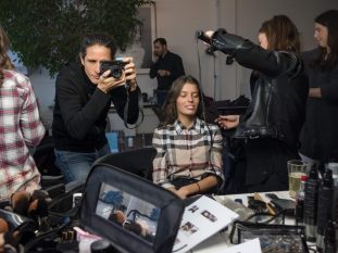 Vue des coulisses d'un shooting de mode montrant un photographe prenant une photo d'un mannequin assis, pendant qu'une coiffeuse s'occupe de ses cheveux. Au premier plan, une table remplie de produits cosmétiques et de pinceaux de maquillage.