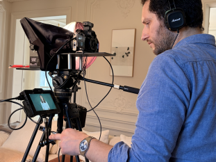 Un photographe professionnel et cadreur de profil portant un casque audio et réglant sa caméra de cinéma avec prompteur installée sur un trépied lors d'une interview.