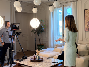 Intervenante en robe blanche debout face à un cadreur et une caméra de cinéma équipée d'un prompteur, lors d'un tournage d'interview dans un salon parisien élégant.